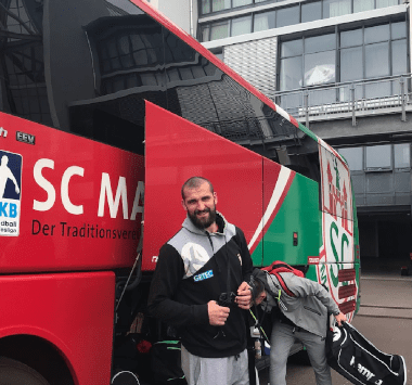 SCM-Trainer Bennet Wiegert steht lächelnd am geöffneten Kofferraum des roten Mannschaftsbusses vor der Abfahrt nach Berlin.