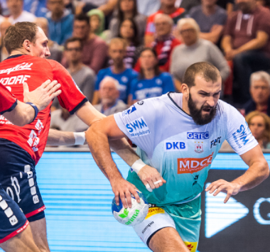 Handball-Actionaufnahme: SC-Magdeburg-Spieler Musa im weißen Trikot spielt den Ball, während ein Verteidiger von SG Flensburg-Handewitt im roten Trikot ihn attackiert, vor Stadionpublikum.