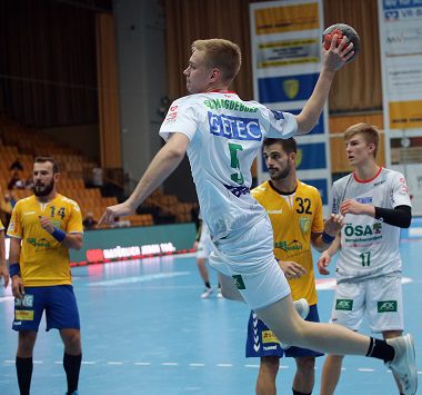 Handball-Actionaufnahme: SC-Magdeburg-Spieler Kay Winter im weißen Trikot springt hoch und wirft den Ball, während Mitspieler und Gegner im Hintergrund zuschauen.