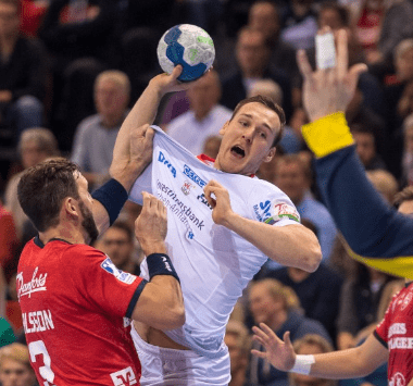 Handball-Actionaufnahme: SC-Magdeburg-Spieler Sulli im weißen Trikot springt hoch und wirft den Ball, während ein Verteidiger von SG Flensburg-Handewitt im roten Trikot ihn attackiert.