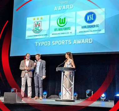 Verleihung des TYPO3 Sports Award: Zwei Männer und eine Frau auf der Bühne vor einer Leinwand mit den Logos von SC Magdeburg, VfL Wolfsburg und Karlsruher SC.