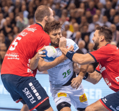 Handball-Actionaufnahme aus dem Spiel SG Flensburg-Handewitt gegen SC Magdeburg: Ein Magdeburger Spieler im weißen Trikot (Nr. 26) behauptet den Ball gegen zwei Verteidiger in roten Trikots.