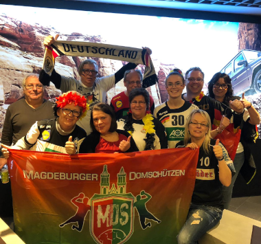 Eine Gruppe von Handballfans in Deutschland-Farben posiert freudig mit der grün-roten Vereinsflagge der „Magdeburger Domschützen MDS“ auf der Tribüne.