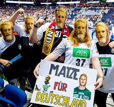 Begeisterte Handballfans auf der Tribüne halten ein selbstgemaltes Schild mit der Aufschrift „MATZE FÜR DEUTSCHLAND“ und einem Porträt von Matthias Musche hoch.