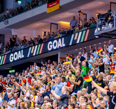Begeisterte Handballfans jubeln mit Deutschland-Fahnen auf der Tribüne unter einer leuchtenden „GOAL“-Bandenwerbung.