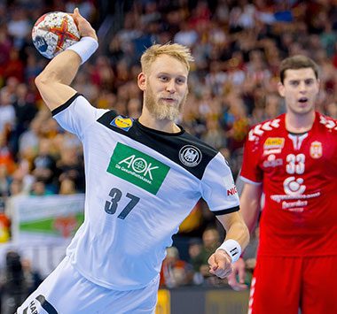 SCM-Linksaußen Matthias Musche (Nummer 37) im weißen Nationaltrikot setzt im Sprungwurf an, während im Hintergrund ein serbischer Spieler im roten Dress zuschaut.