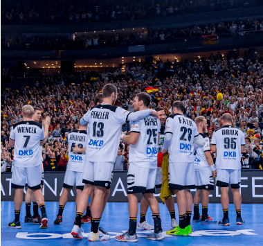 Die deutschen Nationalspieler stehen im weißen DHB-Trikot Arm in Arm auf dem Spielfeld in Köln vor einer vollbesetzten Zuschauertribüne.