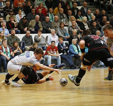 Zwei Handballspieler kämpfen am Boden um den Ball, während ein dritter Spieler herbeiläuft.