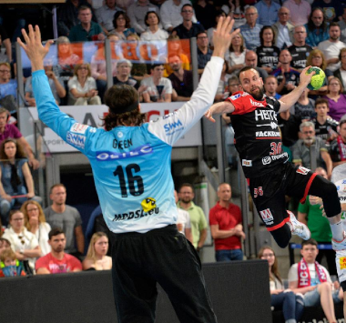 Ein Handballspieler des SC Magdeburg setzt im Sprungwurf zum Torabschluss an, während der gegnerische Torwart die Arme zur Abwehr hochreißt.