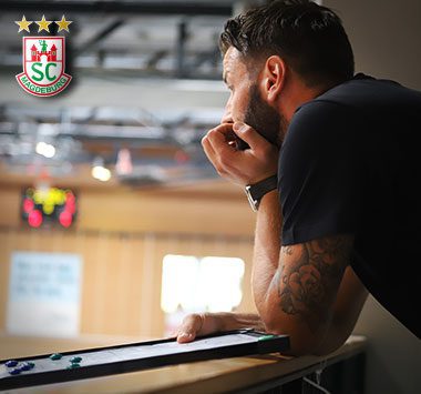 Trainer Bennet Wiegert steht nachdenklich am hölzernen Regiepult am Spielfeldrand und blickt konzentriert in die Handballhalle.