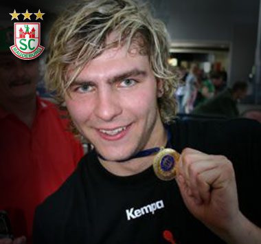 Handballspieler Christoph Theuerkauf im schwarzen Kempa-Shirt blickt lächelnd in die Kamera und hält stolz eine Medaille in der Hand hoch.