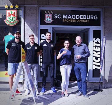 Außenansicht des umgezogenen Fanshops des SC Magdeburg: Offizielle des Vereins präsentieren das neue Ladenlokal mit großem SCM-Logo über dem Eingang.