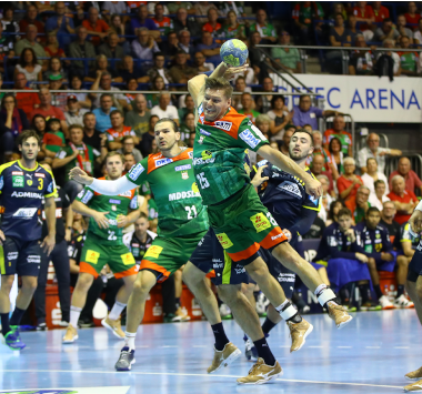 Handballspieler im grün-roten SCM-Trikot steigt im Sprungwurf hoch, während er von der Abwehr der Rhein-Neckar Löwen am Kreis bedrängt wird.