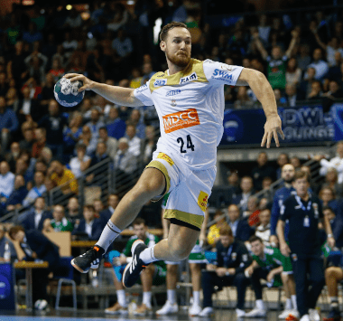 Handballspieler Christian O'Sullivan im weißen SCM-Trikot mit der Nummer 24 springt im Flug weit in den Kreis hinein und setzt zum Sprungwurf an.