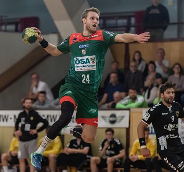 Ein Handballspieler des SC Magdeburg (Nummer 24) springt im grün-roten Trikot hoch in der Luft für einen Wurf, während ein Abwehrspieler der HG Hamburg-Barmbek versucht, ihn zu blocken.
