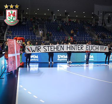 Blick auf das leere Handballspielfeld und die Tribünen; an der Bande hängt ein großes Fan-Banner mit der Aufschrift „WIR STEHEN HINTER EUCH !!!“.