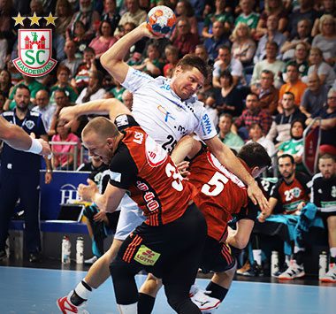 Handballspieler Marko Bezjak springt im weißen Magdeburg-Trikot zum Wurf, während zwei hannoversche Abwehrspieler in Schwarz-Rot ihn attackieren.