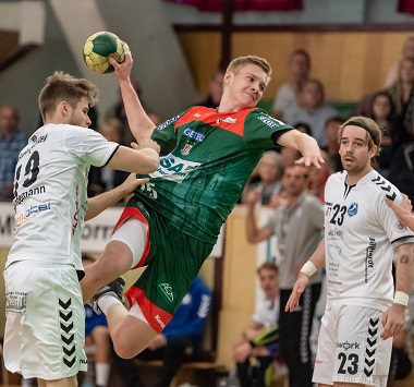Ein Handballspieler des SC Magdeburg II springt im grün-roten Trikot zum Wurf hoch, während zwei weiße Abwehrspieler des HSV Hannover versuchen, ihn zu blocken.