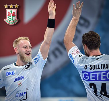 Die Handballspieler Matthias Musche und Christian O'Sullivan klatschen im weißen Magdeburg-Trikot freudig auf dem Spielfeld ab.