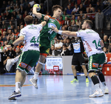 Handballspieler Michael Damgaard springt im grün-roten Trikot des SC Magdeburg zum Wurf hoch, während zwei Göppinger Abwehrspieler in Weiß-Grün ihn blocken.
