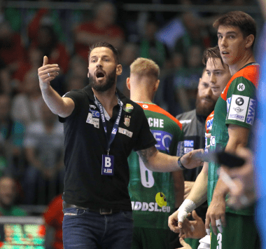 Handball-Trainer Bennet Wiegert gibt am Spielfeldrand lautstark Anweisungen; im Hintergrund stehen Magdeburger Spieler im grün-roten Trikot.