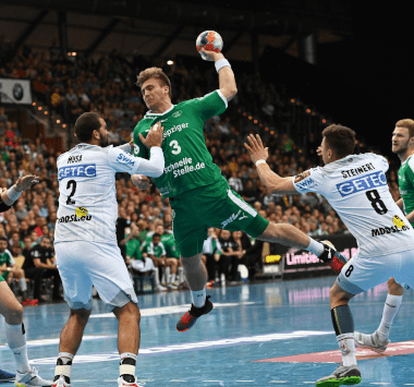 Handballspieler Albin Lagergren springt im grünen Magdeburg-Trikot (Nummer 3) hoch zum Wurf, während ihn drei Leipziger Abwehrspieler in Weiß bedrängen.