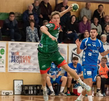 Ein Handballspieler des SC Magdeburg II springt im grün-roten Trikot zum Wurf hoch, während ein Potsdamer Abwehrspieler in Blau die Bewegung verfolgt.