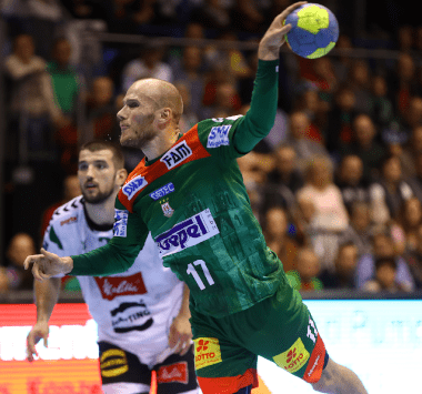 Handballspieler Daniel Pettersson springt im grün-roten Magdeburg-Trikot zum Wurf hoch, während ein Mindener Abwehrspieler in Weiß-Schwarz hinterherläuft.