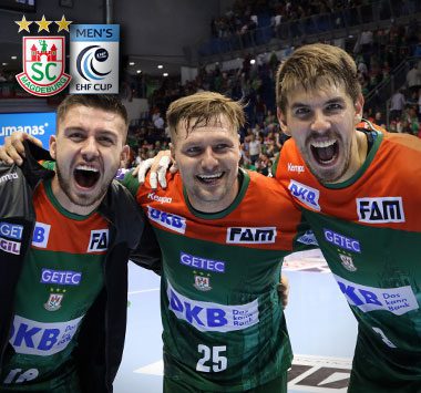 Drei Handballspieler des SC Magdeburg, darunter Marko Bezjak (Mitte, Nummer 25), jubeln im grün-roten Trikot Arm in Arm auf dem Spielfeld.