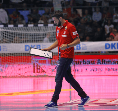 Handball-Trainer Bennet Wiegert läuft im roten Poloshirt am Spielfeldrand entlang und hält eine Taktiktafel unter dem Arm.