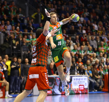 Handballspieler Albin Lagergren springt im grün-roten Trikot des SC Magdeburg (Nummer 23) zum Wurf hoch, während ein Abwehrspieler der Füchse Berlin in Rot ihn blockt.