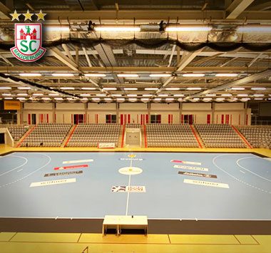 Blick von den Rängen auf das leere hellblaue Spielfeld einer Handballhalle (HUK-COBURG arena) mit aufgebauten Werbebanden vor einem Geisterspiel.
