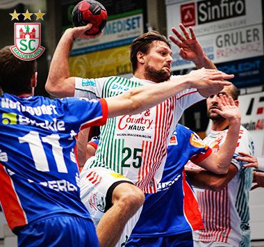 Handball-Spielszene: SCM-Rückraumspieler Marko Bezjak (Nummer 25) holt im weiß-rot gestreiften Auswärtstrikot zum Wurf aus, während er von zwei Balinger Abwehrspielern in blauen Trikots bedrängt wird.