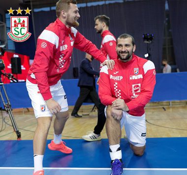 Nationalmannschafts-Szene: Der kroatische Nationalspieler Željko Musa kniet lächelnd im roten Trikot auf dem blauen Spielfeld, während ein Teamkollege im weißen Trainingsanzug hinter ihm steht.