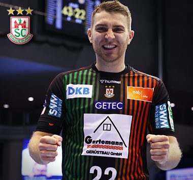 Spielerporträt: SCM-Linksaußen Lukas Mertens blickt im grün-schwarz gestreiften Heimtrikot (Nummer 22) lächelnd frontal in die Kamera.