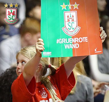 Ein junges Mädchen im roten SCM-Shirt hält auf der Zuschauertribüne ein grün-rotes Pappschild mit dem Hashtag „#GRUENROTE WAND“ hoch.