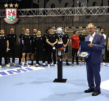 Ehrung auf dem Spielfeld: Ministerpräsident Reiner Haseloff steht im blauen Anzug mit einer Urkunde auf der Platte, während im Hintergrund das Handball-Team des SC Magdeburg applaudiert.