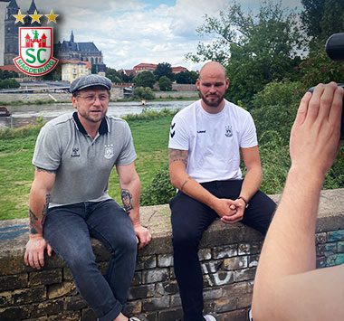 Torhüter Mike Jensen sitzt im weißen SCM-Shirt neben einem Interviewer auf einer Steinmauer an der Elbe und blickt in die Kamera.