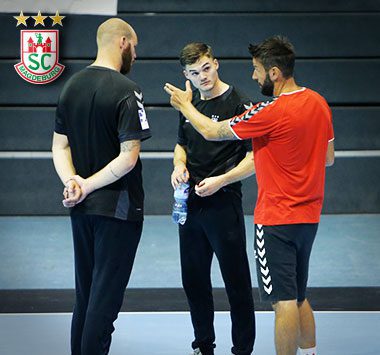 Zwei SCM-Spieler und Trainer in Trainingskleidung stehen in der Halle zusammen und unterhalten sich beim offiziellen Trainingsstart.
