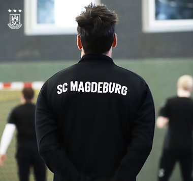 Rückansicht eines Spielers in einem schwarzen Trainingspullover mit der Aufschrift „SC Magdeburg“ auf dem Spielfeld.