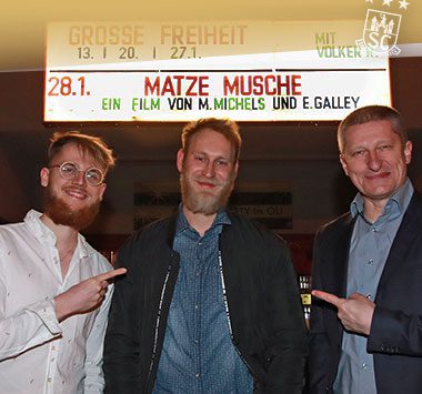 Matthias Musche steht lächelnd mit zwei Männern vor dem Filmplakat seiner Kinodokumentation