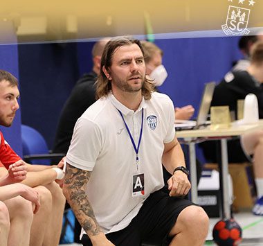 Christoph Theuerkauf sitzt im weißen Trainer-Shirt fokussiert an der Seitenlinie.