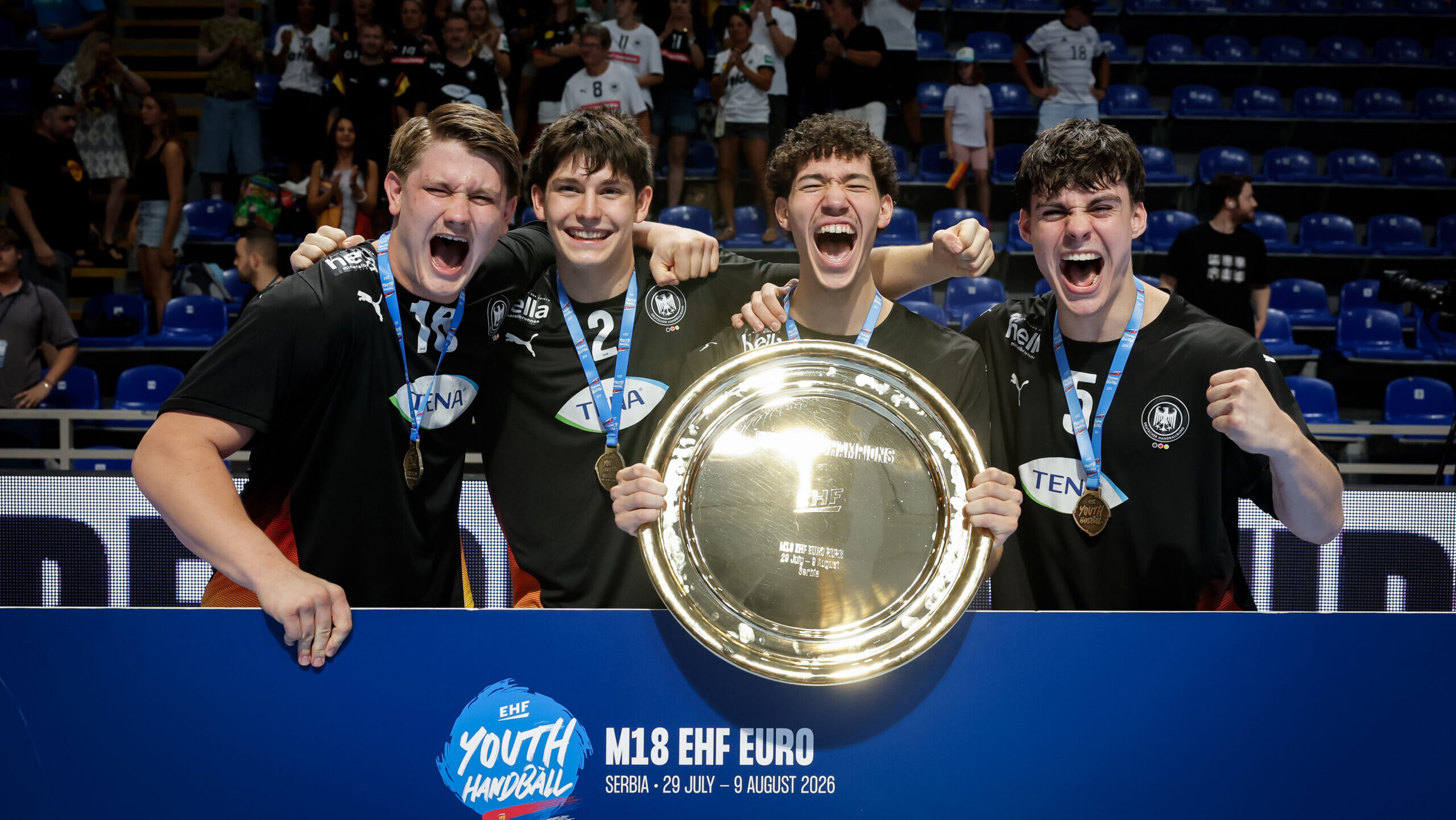 Four athletes in black jerseys celebrate with a large golden trophy, wearing medals and cheering enthusiastically at the M18 EHF Euro event.