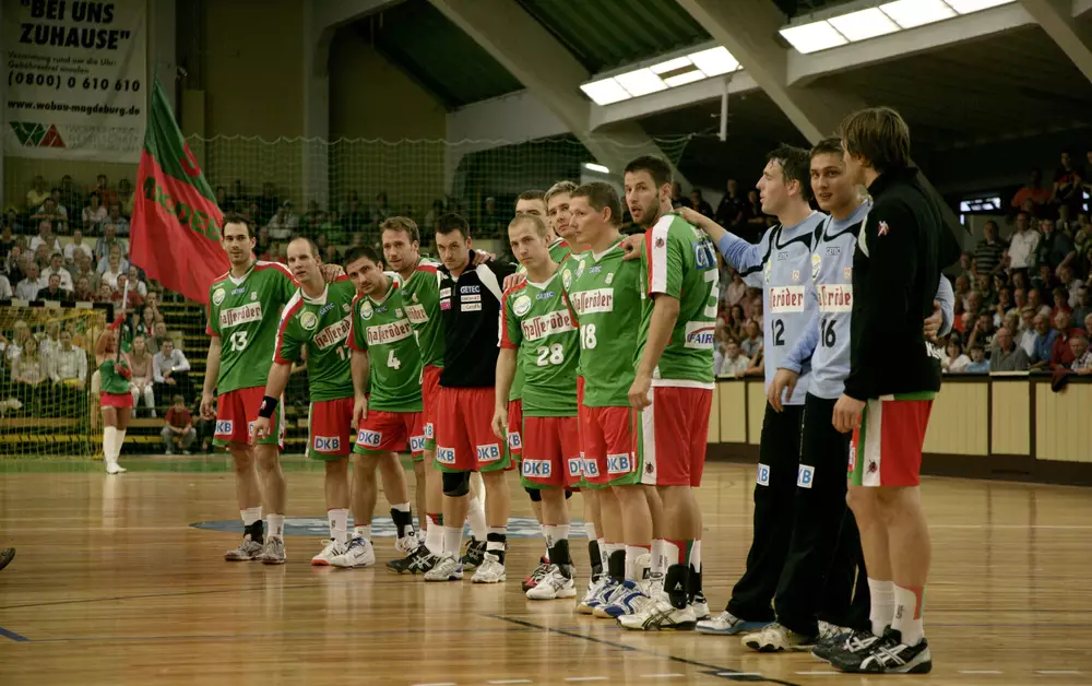 Spieler des SC Magdeburg, darunter Bennet Wiegert (Nummer 13), stehen nebeneinander auf dem Spielfeld in der Bördelandhalle.